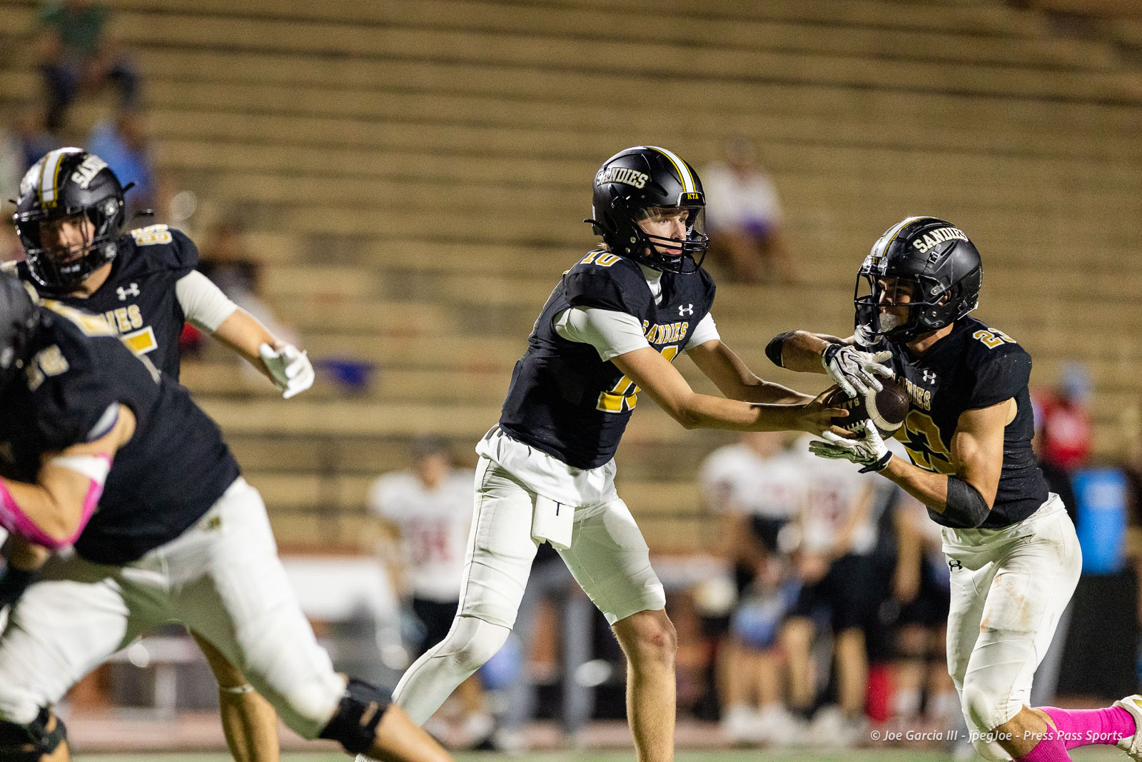lubbock-monterey-vs-amarillo-high-football-jpegJoe-pps-34 | Press Pass Sports Football players in black-and-gold uniforms scramble for the ball during a night game, with the center reaching as a teammate grips the ball.