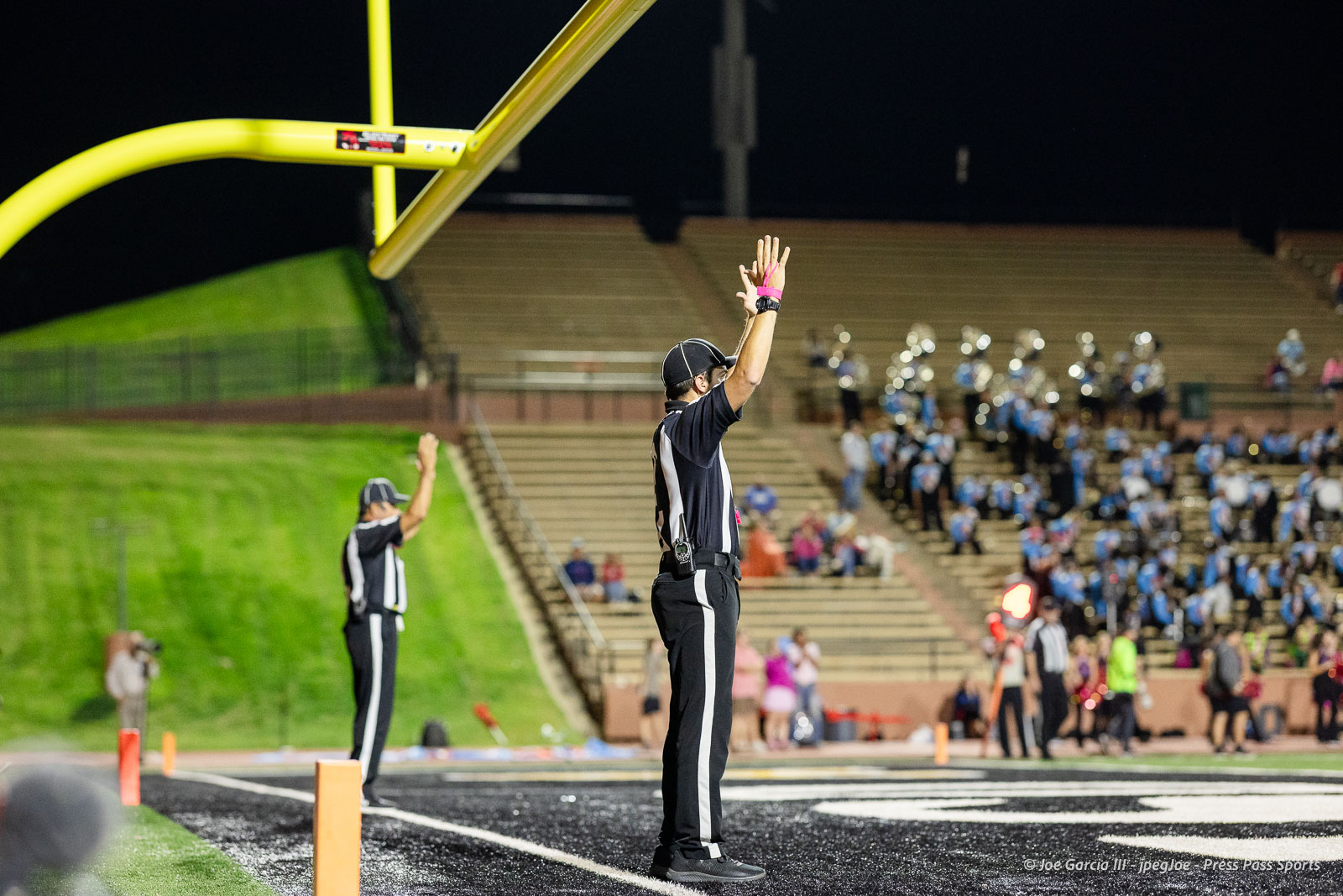 Football referee on the sideline signaling a touchdown with both arms raised during a night game, stadium lights in the background.]","Official in black and white raises both arms to signal a score, stands near the end zone as a crowd watches.]","Referee signaling touchdown at a night football game, yellow goalposts and stands visible behind him.]","Upright referee signals a score near the end zone while players and spectators look on under stadium lights.]","Night football game: referee raises arms in triumph as the crowd watches from the stands near the end zone.