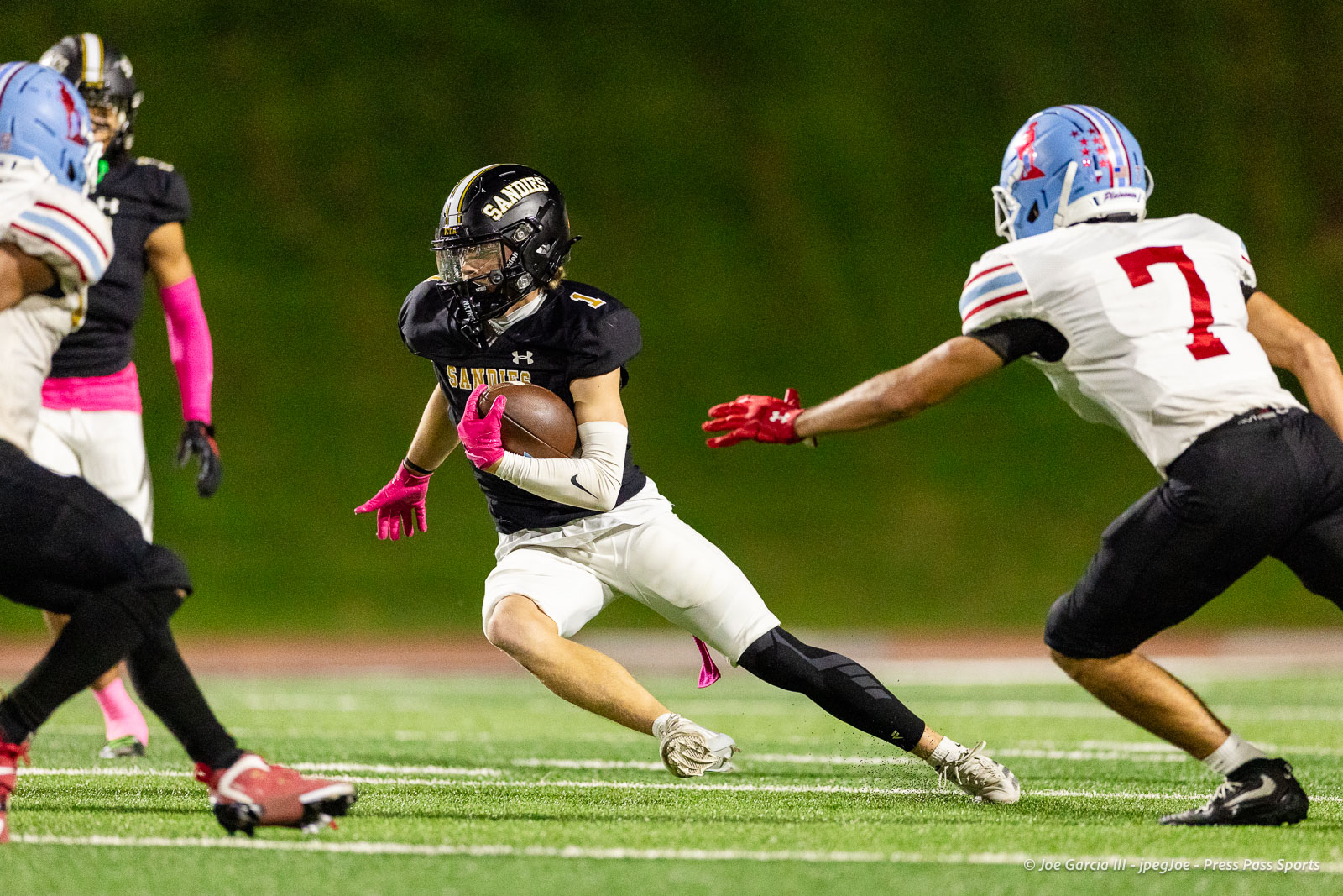 lubbock-monterey-vs-amarillo-high-football-jpegJoe-pps-37 | Press Pass Sports Football: a running back in a black Sandies jersey with pink gloves carries the ball while dodging a defender in white (number 7) on a green turf field at night.