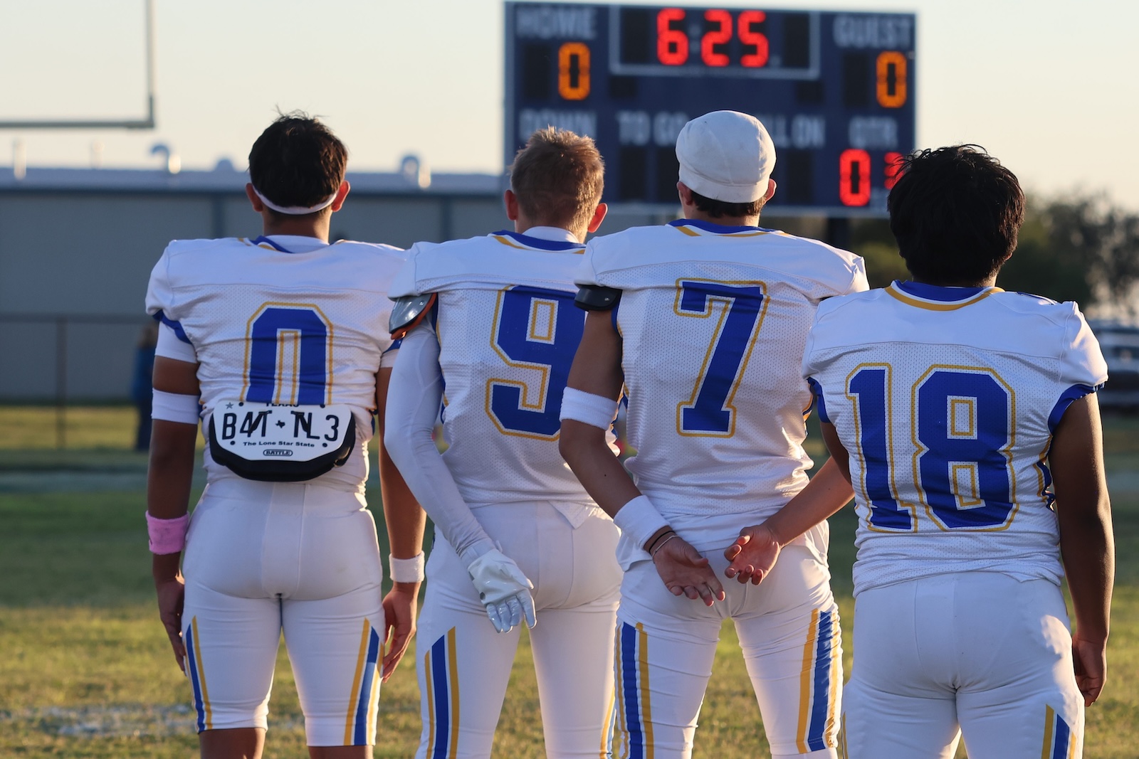 Four football players in white and blue uniforms stand with their backs to the camera on a grassy field, a scoreboard visible in the distance.