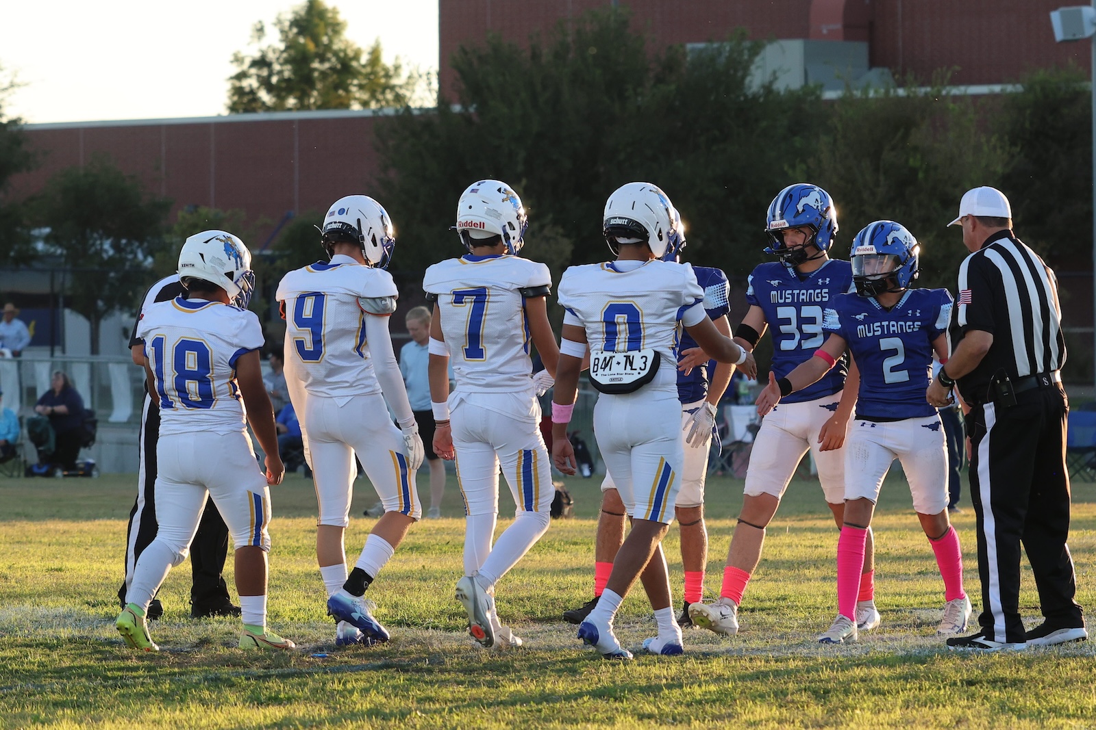 Youth football players in white and blue uniforms form a circle on a grassy field as a referee watches, pre-game or during a break.