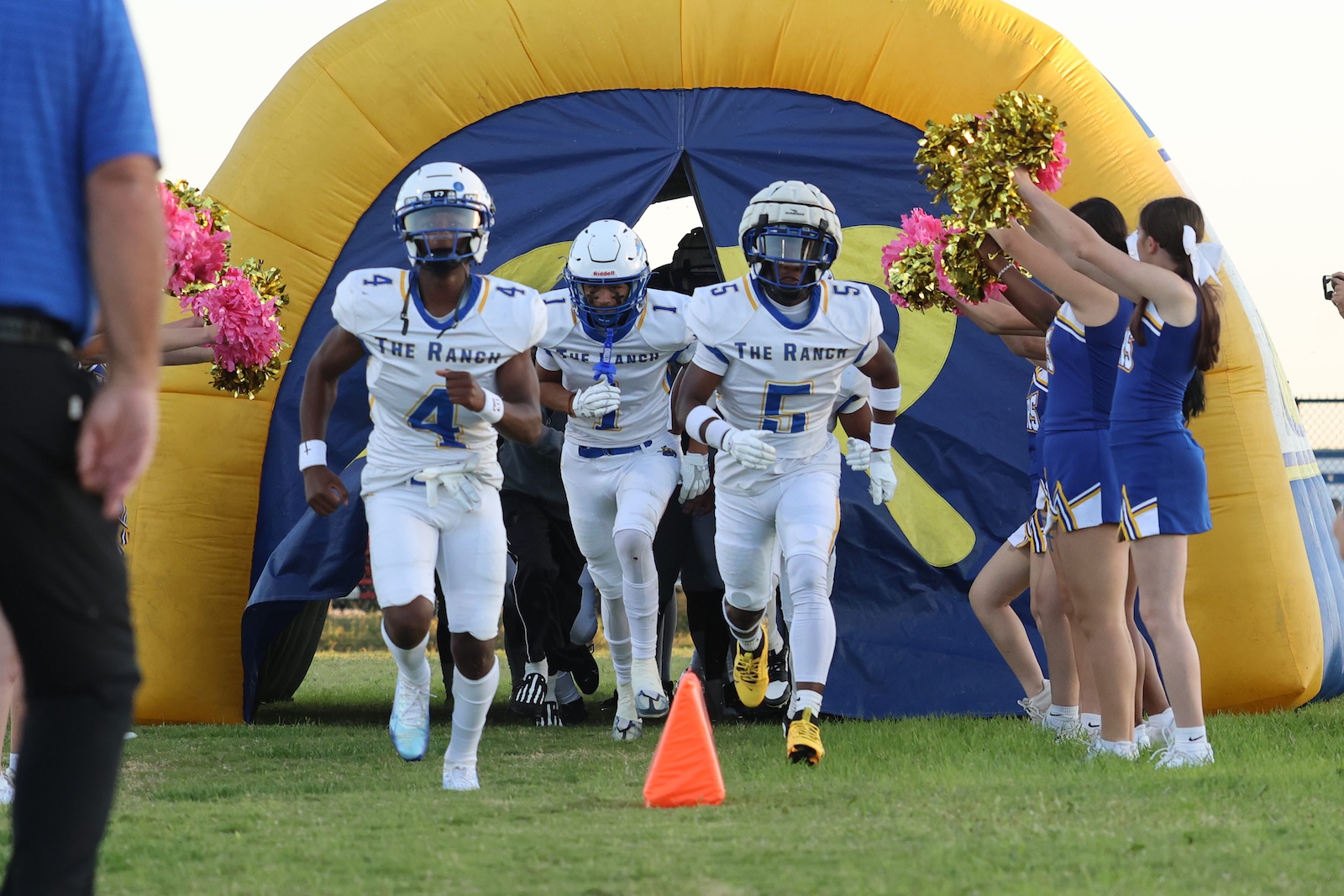 Football players from The Ranch sprinting onto the field through a blue-yellow inflatable tunnel, cheerleaders on the sides holding pom-poms in blue uniforms with pink accents.