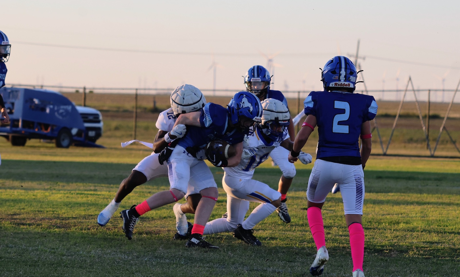 Youth football game: a blue team player tackled by white-team players as he carries the ball on a grassy field during sunset/silver hour.