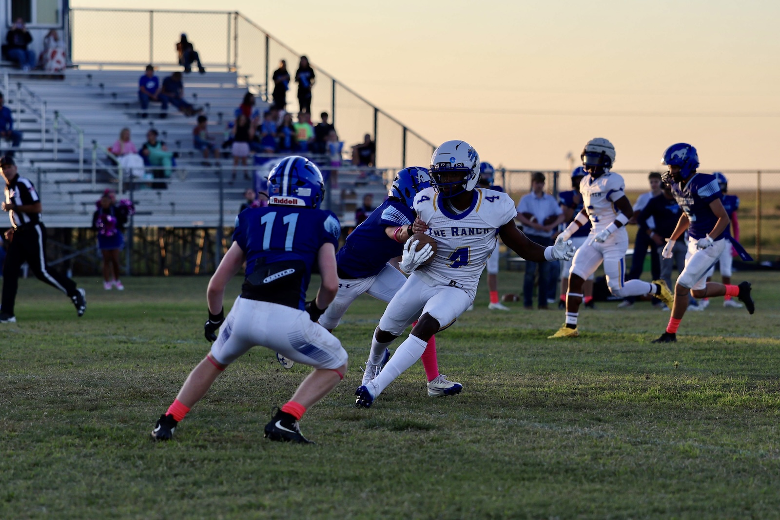 White-uniform running back with the ball dodges blue defenders on a grassy field as bleachers and spectators loom in the background at sunset.