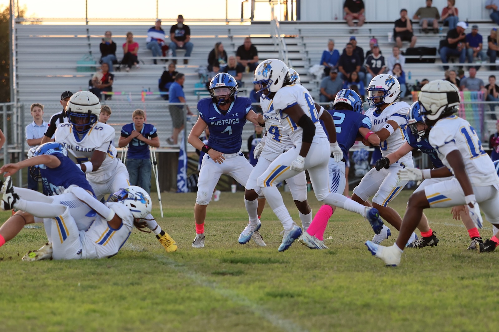High school football game action: blue Mustangs players tackle against white-uniformed Ranch players on a grassy field, with stands in background.