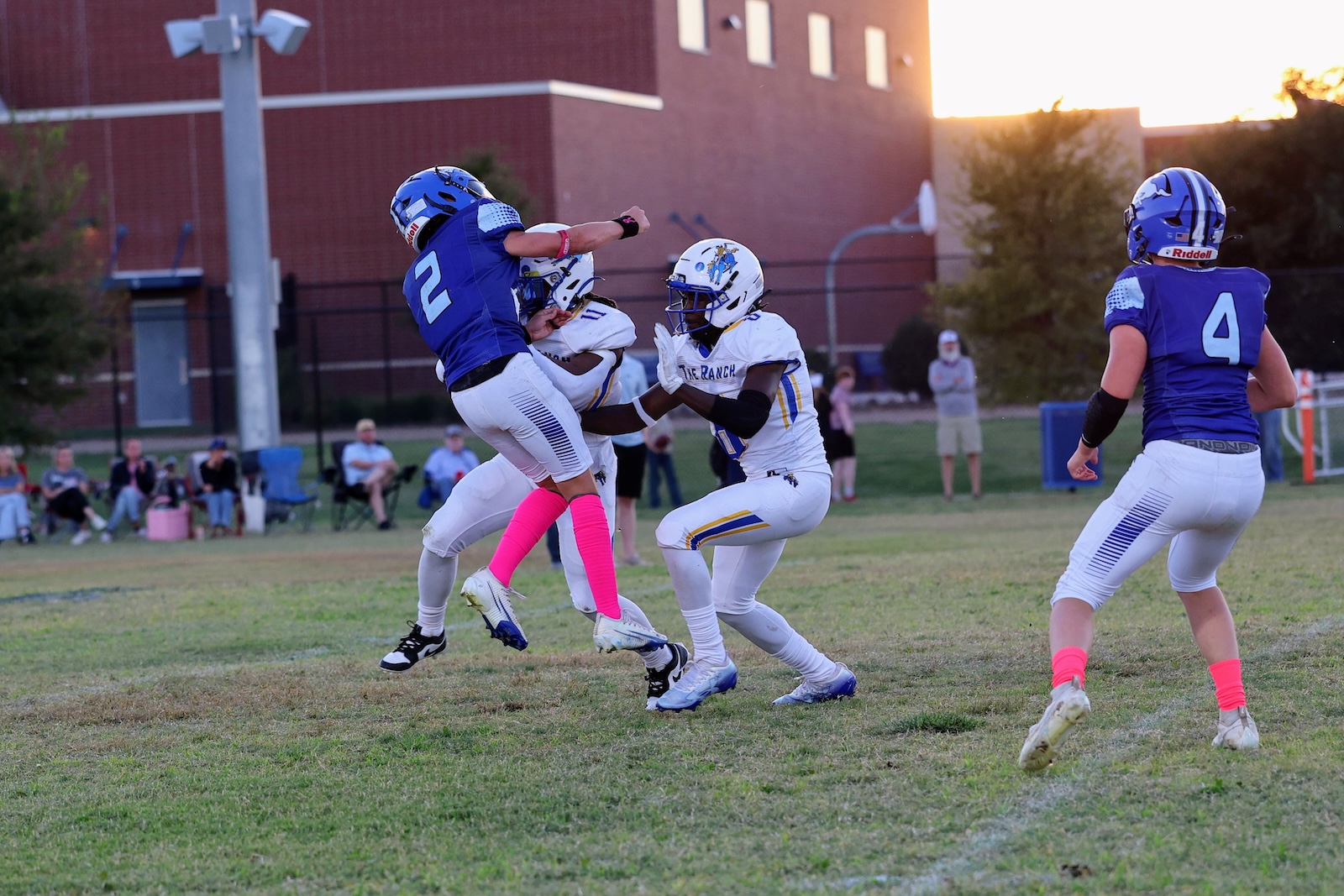 Football game action: a blue-team defender jumps to tackle a white-team player on a grassy field as others watch in the background.