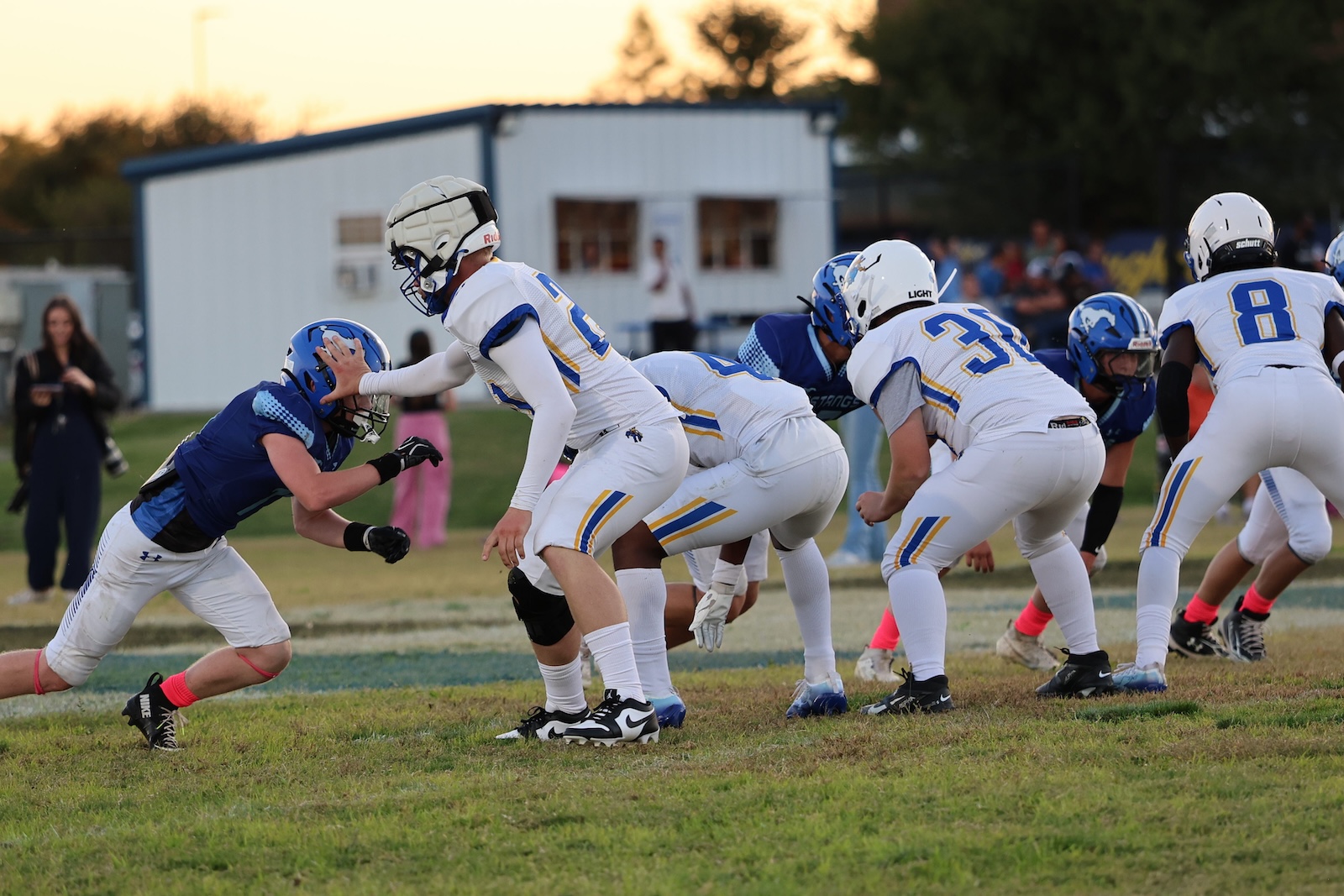 Youth football players in white and blue line up at the scrimmage on a sunlit field.