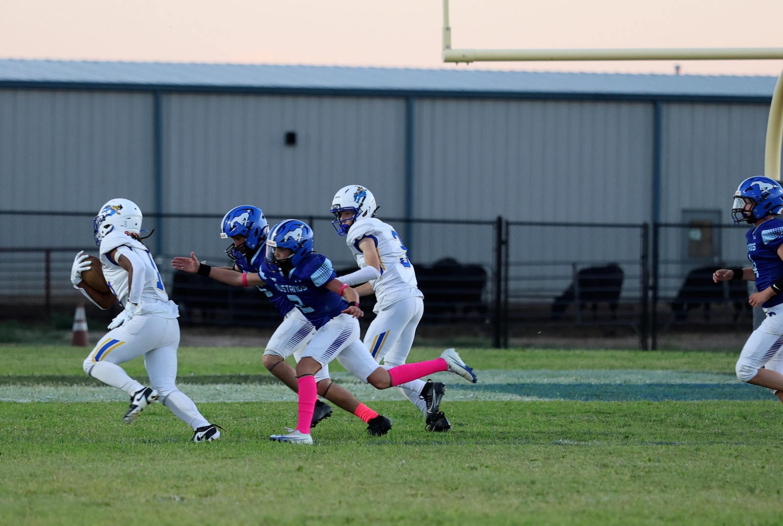 Youth football players in white and blue uniforms chase the ball on a grassy field during a game, one player carrying the ball as others pursue.