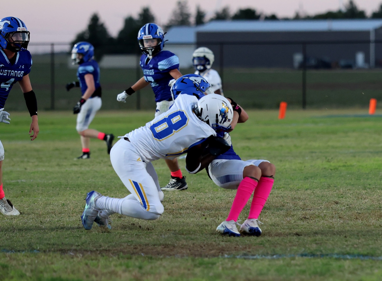 White-uniformed player wearing number 68 tackles a blue-and-white opponent on a grassy football field during a game; teammates in blue jerseys watch in the background, pink socks visible on another player, with orange pylons in the distance.