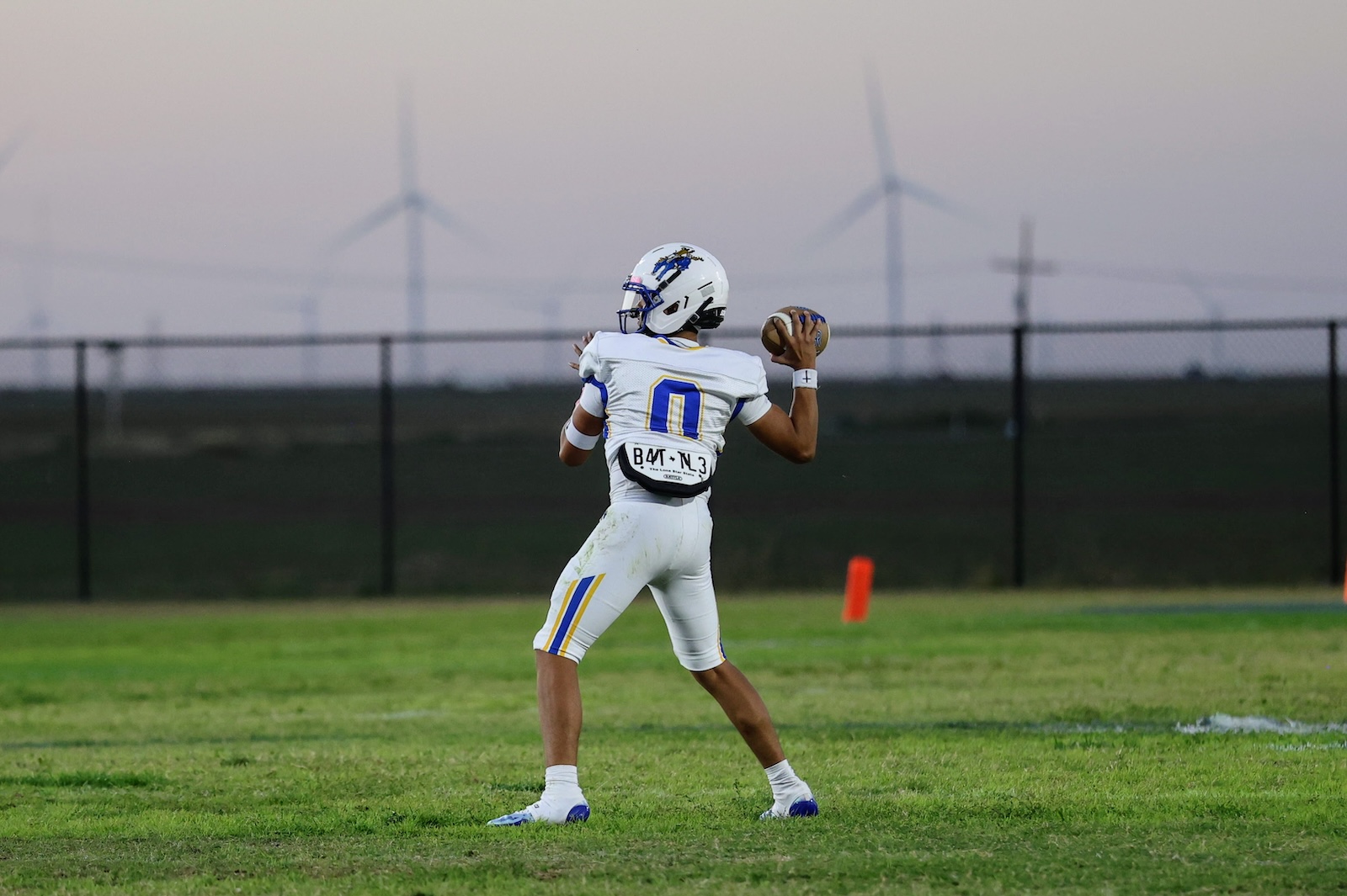 Quarterback in white jersey #0 preparing to throw a pass on a grassy field with a fence and distant wind turbines in the background.