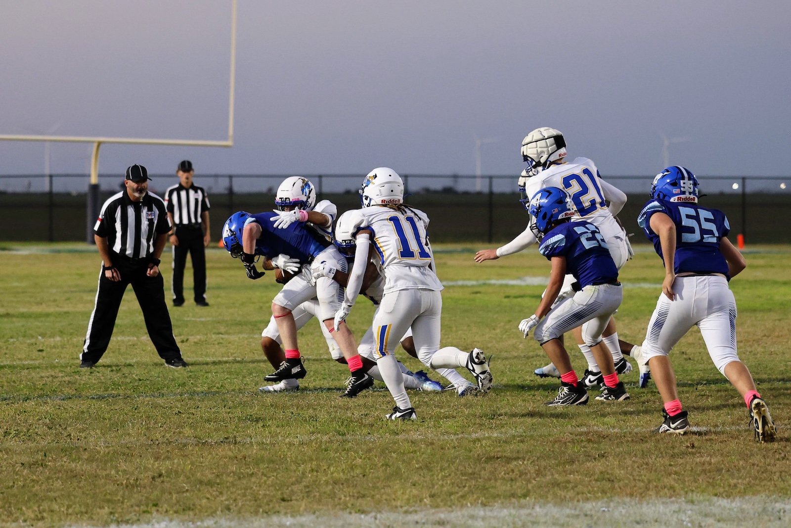 Football players in white and blue jerseys clash as a tackle occurs near the line of scrimmage on a grassy field at dusk.