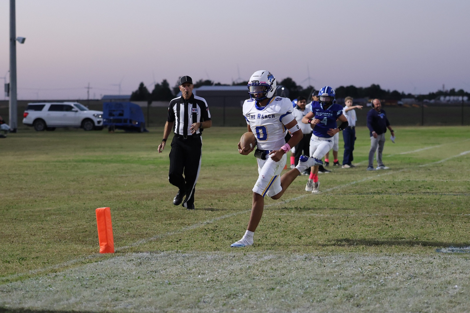 Football player in white-and-blue uniform runs with the ball toward the end zone, near an orange pylon on a grassy field as a referee watches nearby.