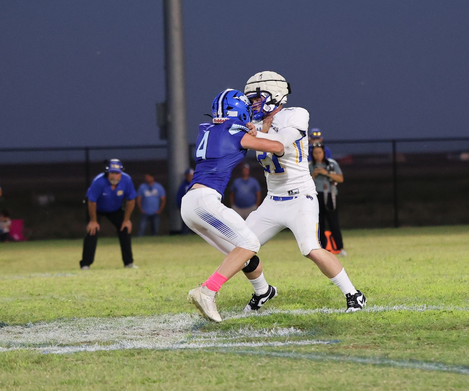 Two football players collide on a grassy field at dusk, blue jersey #4 and white jersey #11 grappling for control.