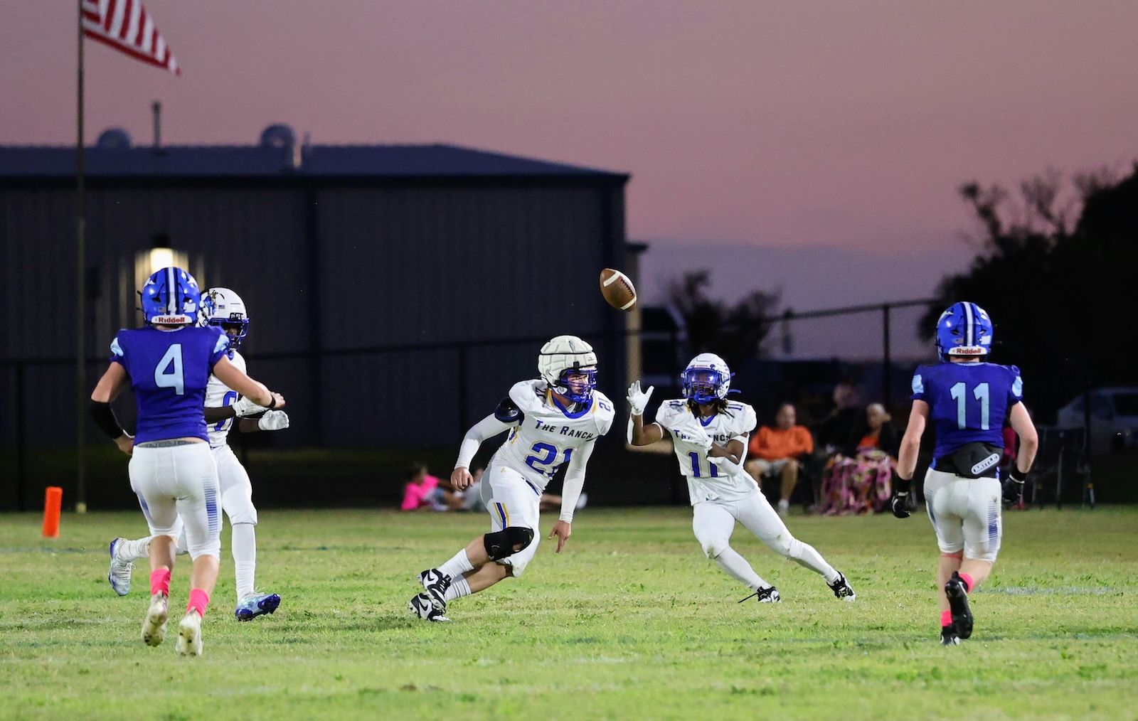 Two white-uniform players (#21 and #11) reach for a football in mid-air as blue-team defenders close in during a dusk game.