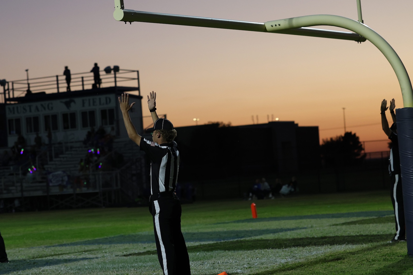 Football referee signals a touchdown with arms raised on a high school field at sunset, Mustang Field in the background.