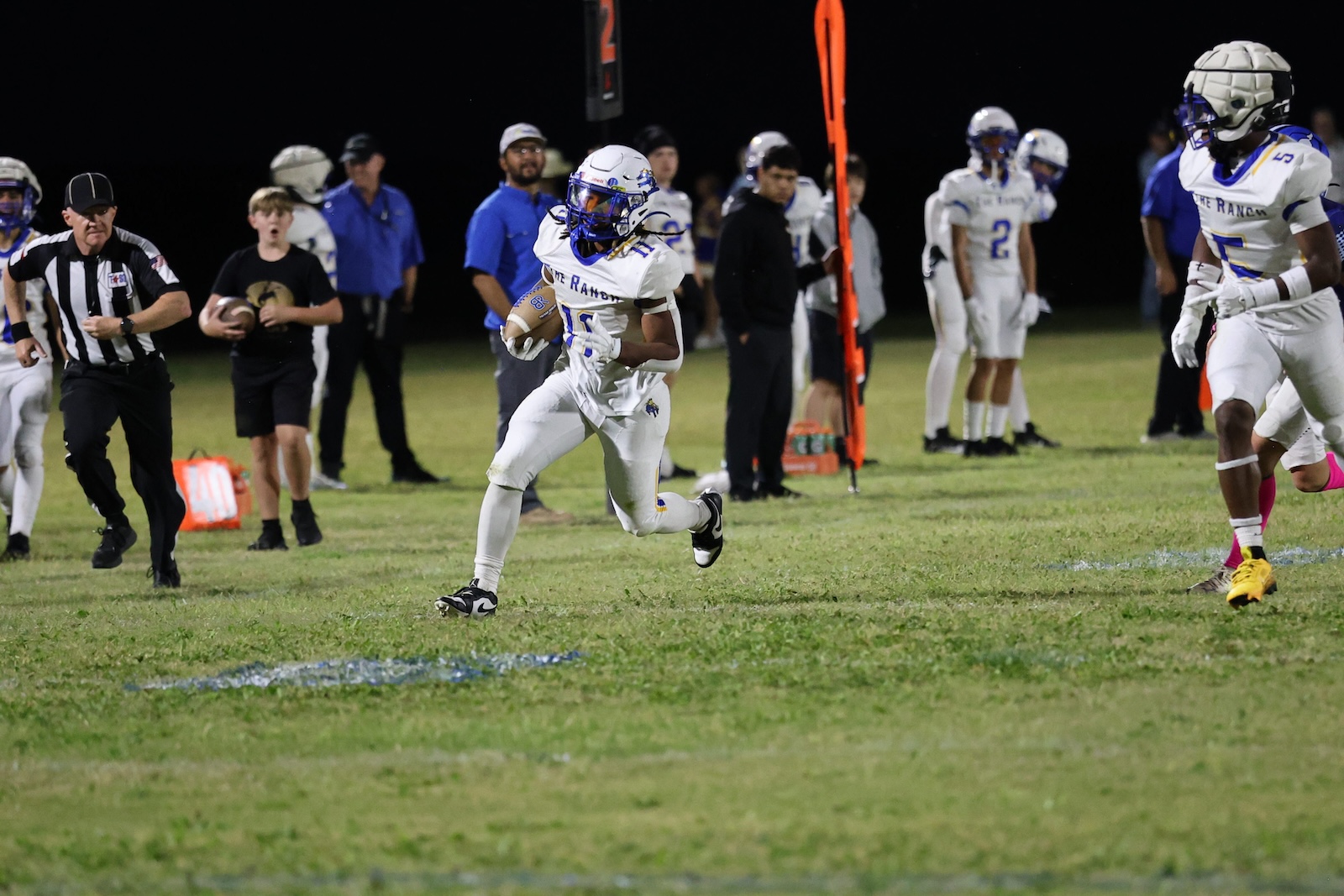 Football player in white uniform carries the ball downfield during a night game, with teammates and officials in the background.