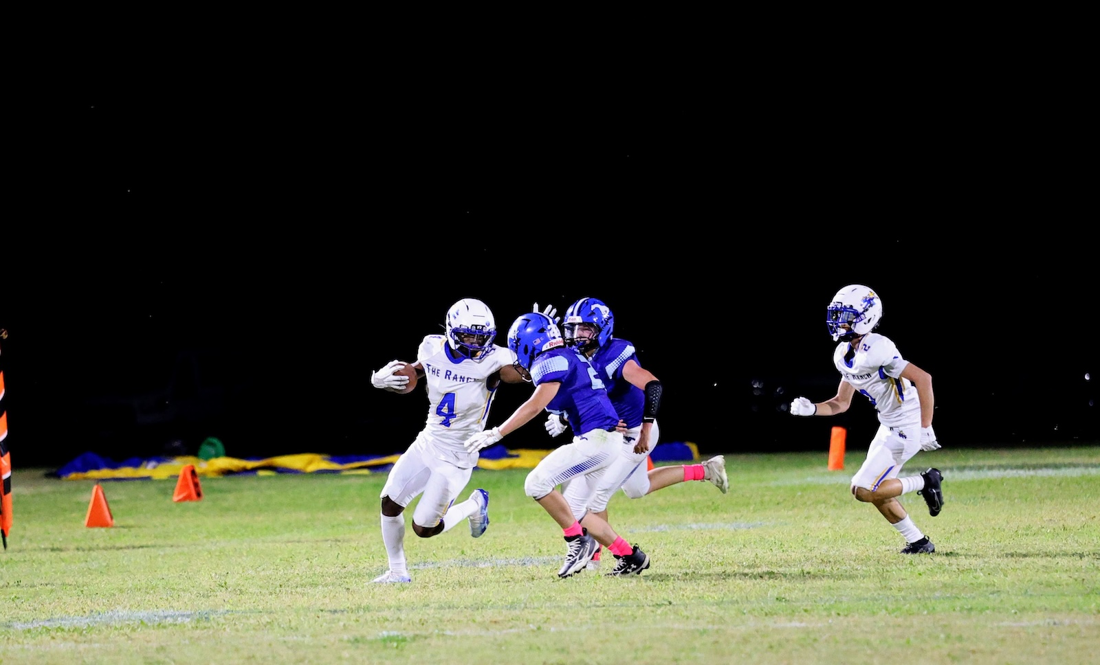 Night-time football scene: a white-team player wearing number 4 runs with the ball as blue-uniform defenders chase on a lit field with orange pylons nearby.