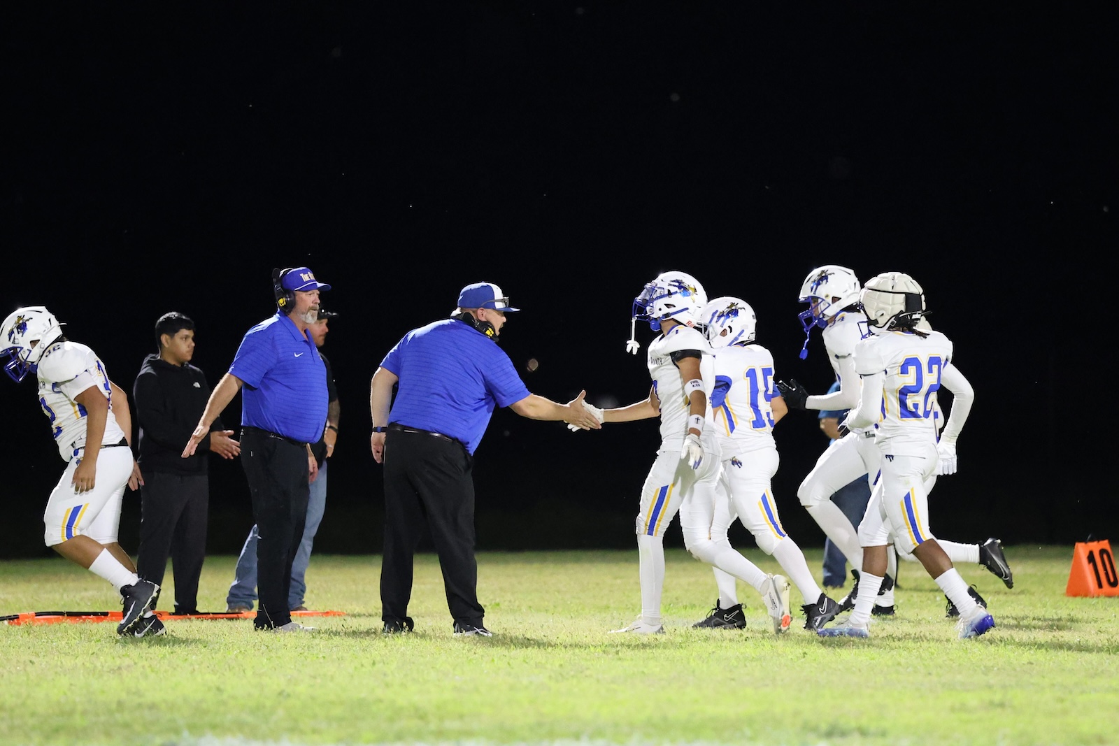 Coach in blue shirt shakes hands with a football player during a night game on a grassy field.