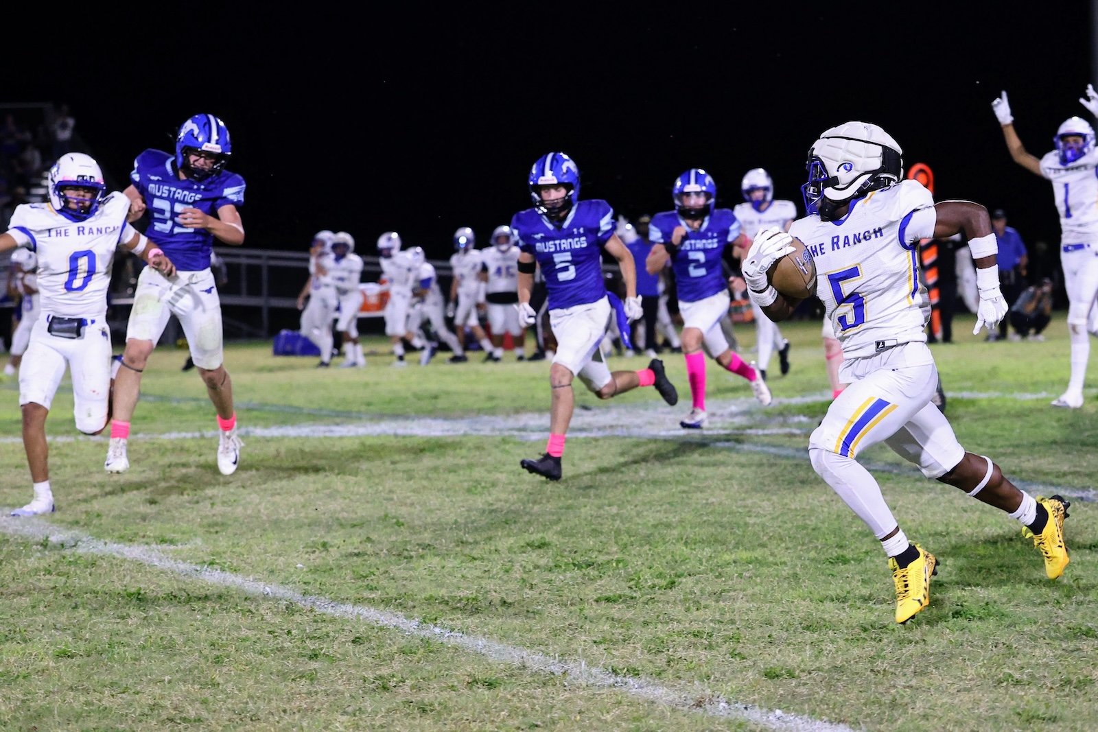 A football player in white The Ranch jersey (#5) runs with the ball while teammates and defenders pursue at night on a grassy field.
