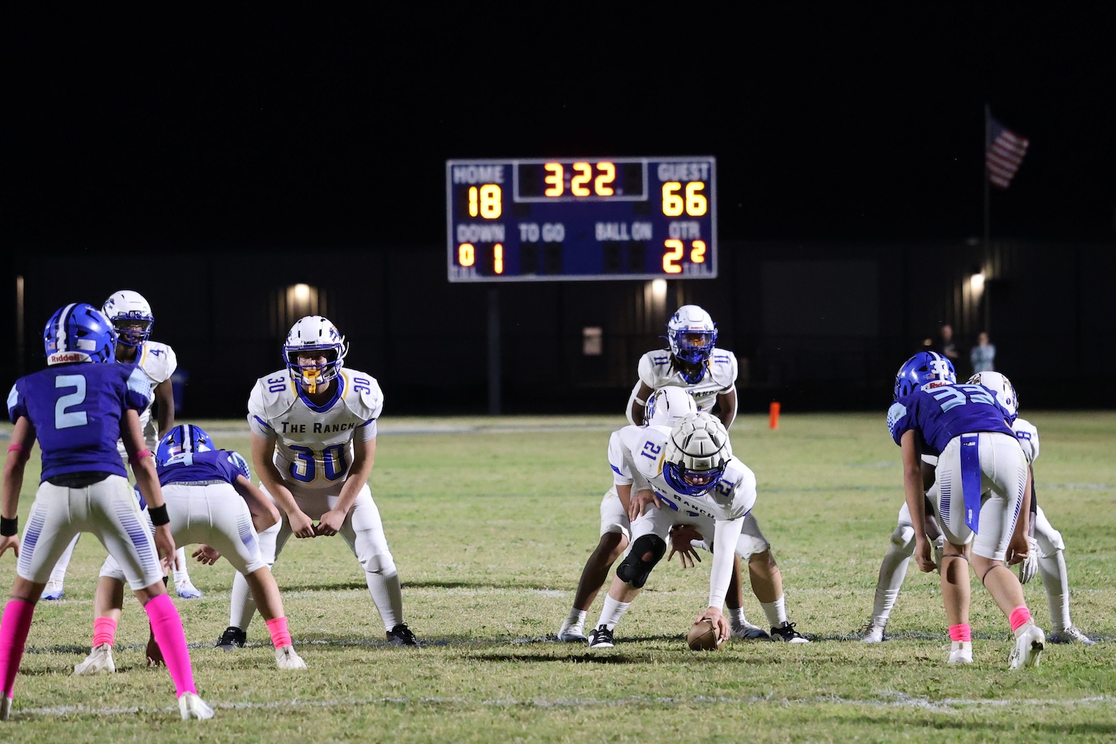 Night football game: linemen line up at the snap with a bright score board in the background showing the teams’ points, 18–66 edge labeled HOME/GUEST.