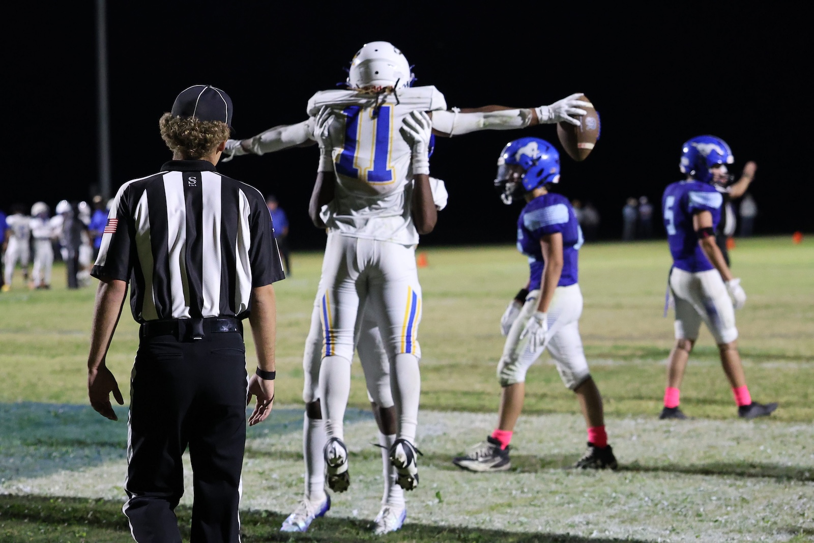 Nighttime football scene: a player in white and blue is lifted by a teammate while a referee watches from the foreground.