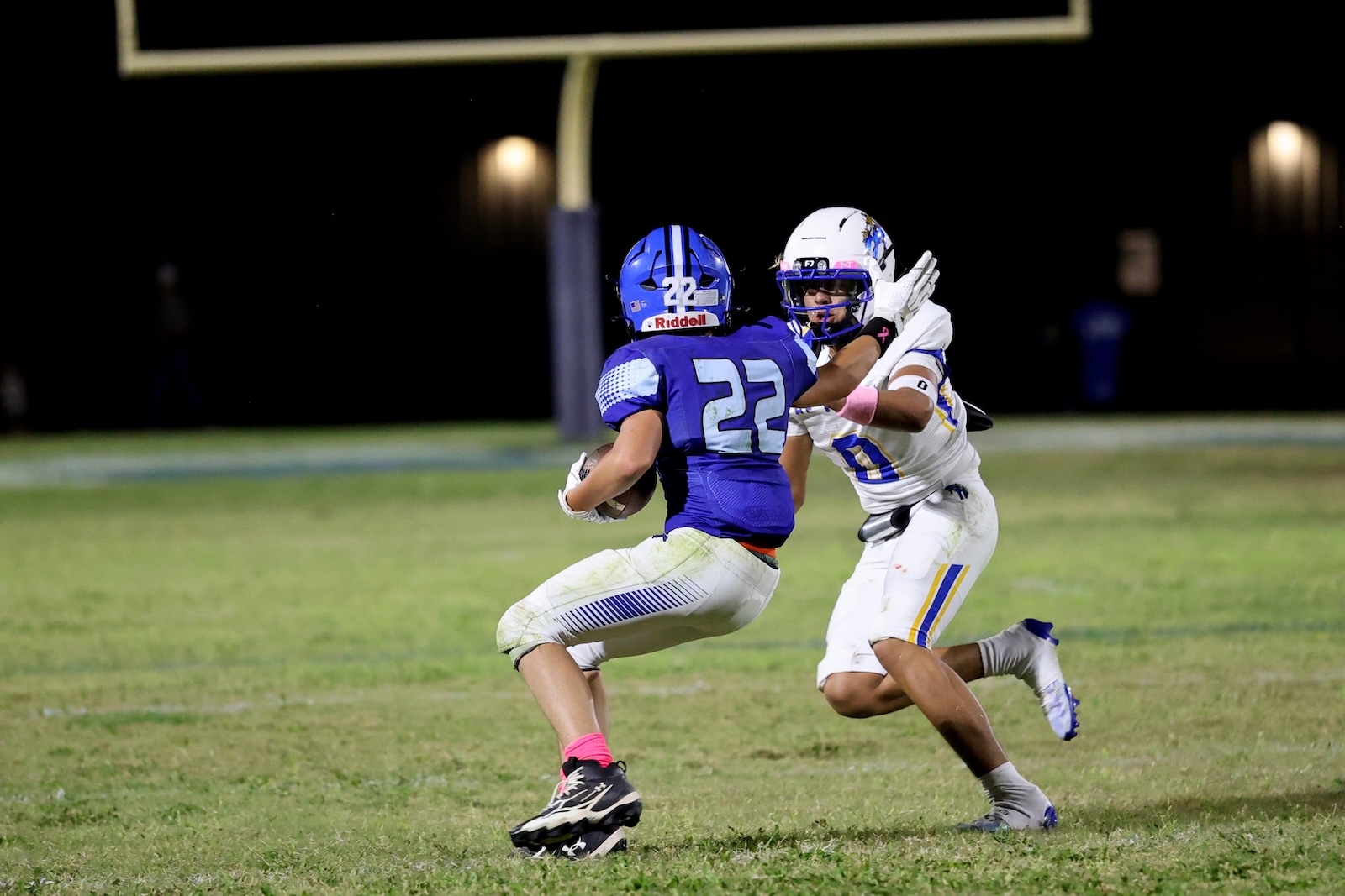 Football players collide on a night game; blue jersey #22 carries the ball while a defender in white reaches in for a tackle, goalposts in background
