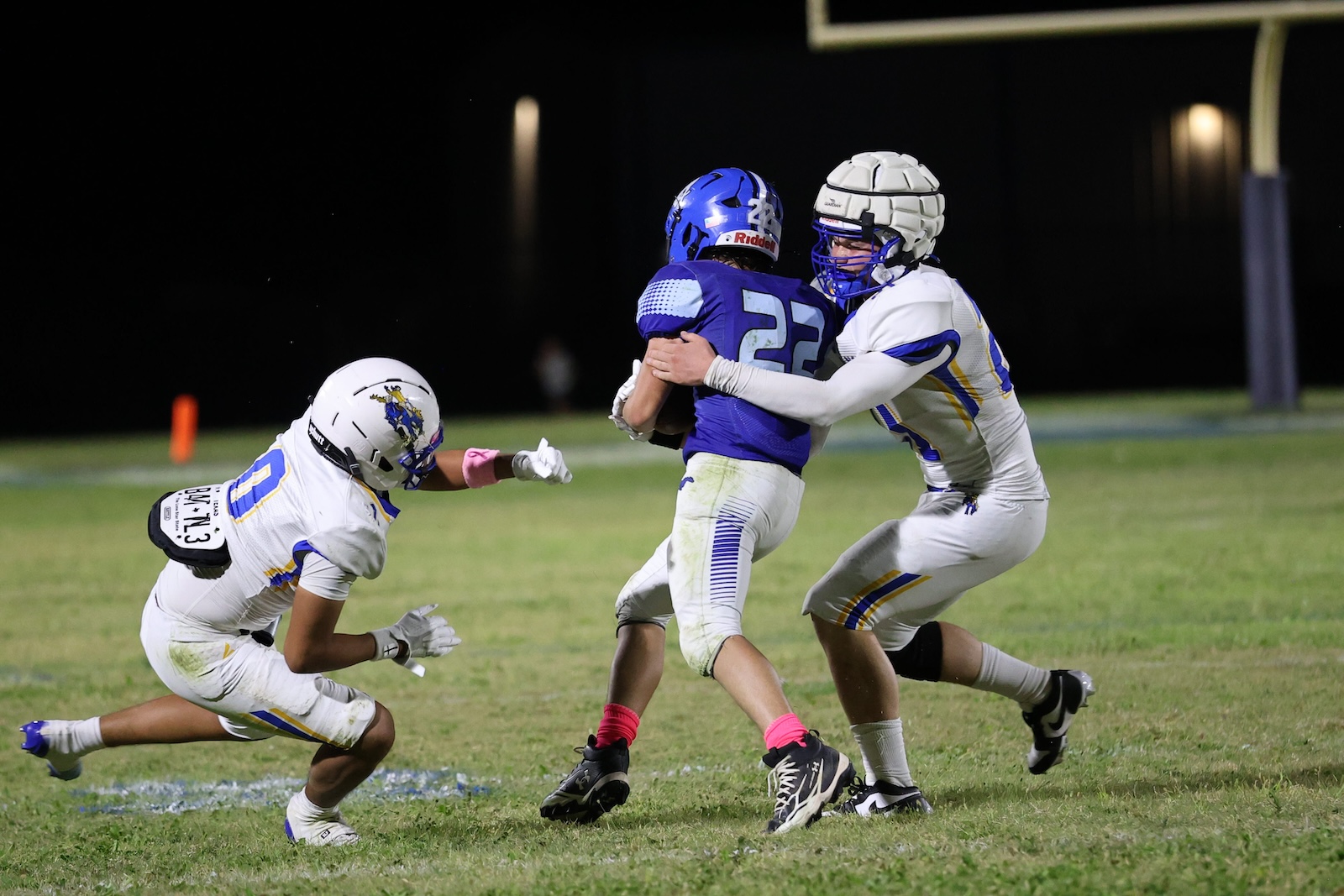Nighttime football action: a white-helmet defender tackles a blue jersey runner as another white-jersey player closes in on the play.