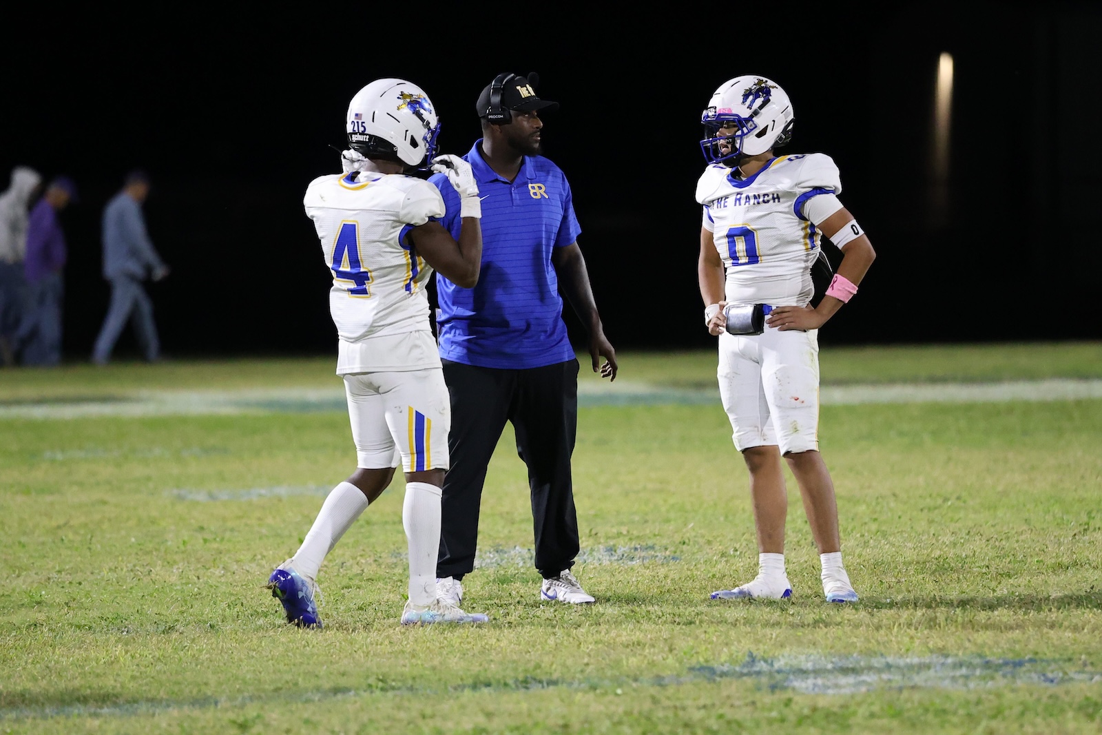 Two football players in white uniforms with blue and gold trim stand with a coach in a blue polo on a grassy field at night, discussing strategy.