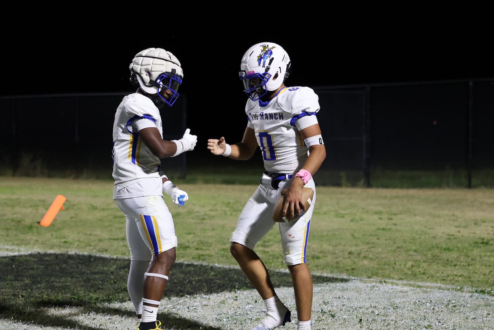 Two high school football players in white jerseys share a fist bump on a night game, one holding a football on the field