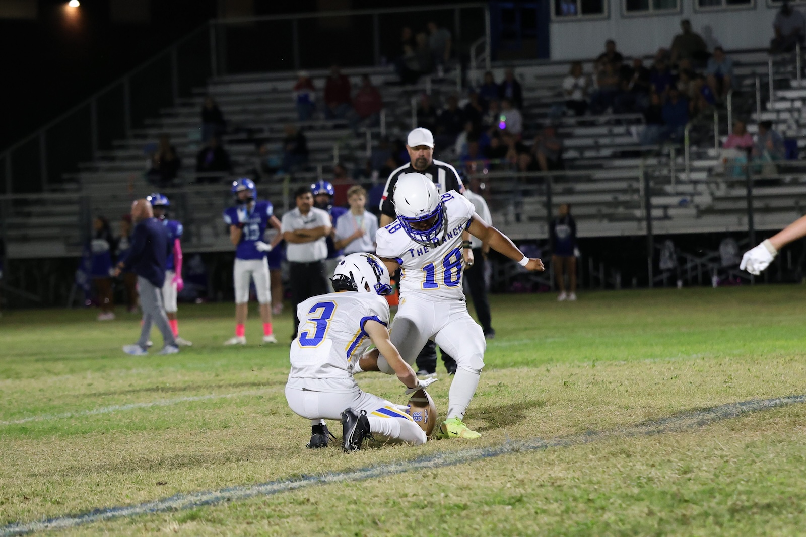 Football players in white and blue uniforms on a night game; one kneels to hold the ball while another runner (#18) approaches for a play, with bleachers in the background.