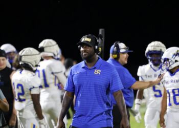 PY8A8892 | Press Pass Sports Coach in a blue polo with a headset walking along the sideline as players in white uniforms stand behind him on the football field at night.