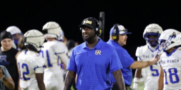 Coach in a blue polo with a headset walking along the sideline as players in white uniforms stand behind him on the football field at night.