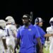 PY8A8892 | Press Pass Sports Coach in a blue polo with a headset walking along the sideline as players in white uniforms stand behind him on the football field at night.