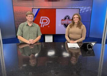 IMG_8100 | Press Pass Sports Two news anchors sit at a glossy studio desk in front of a large screen displaying red and purple panels with logos behind them, smiling at the camera.