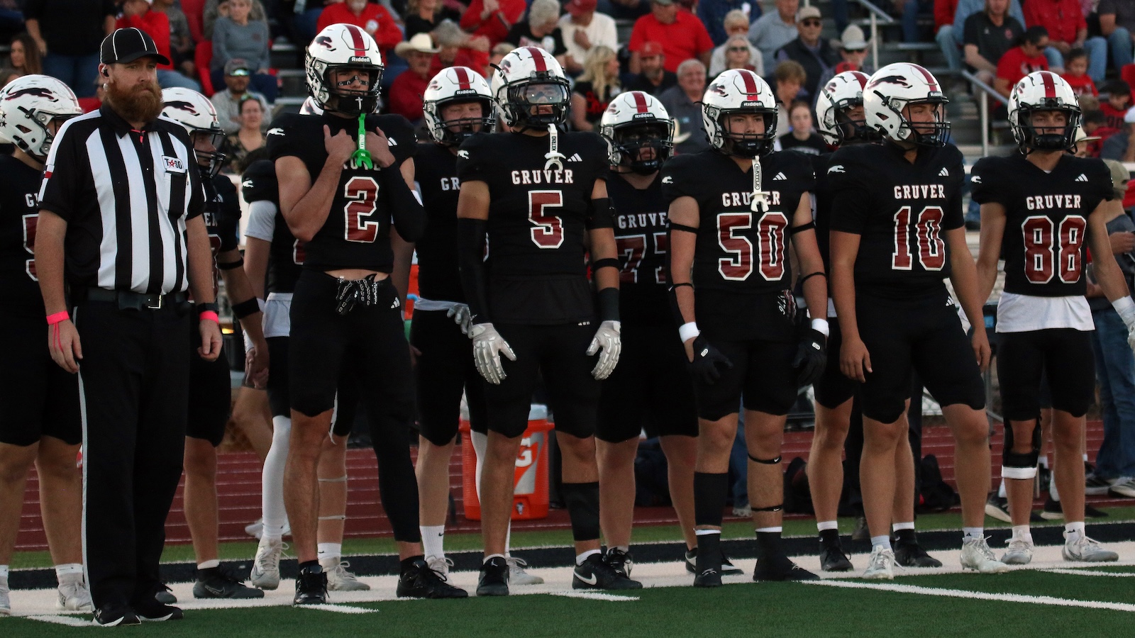 A high school football team in black uniforms lined up on the sideline with a referee on the left and a crowd in the stands behind them.