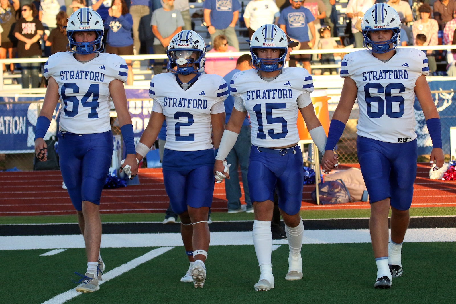 Four Stratford football players in white jerseys with blue stripes walk hand in hand onto the field, numbers 64, 2, 15, and 66, with a cheering crowd in the stands behind them.