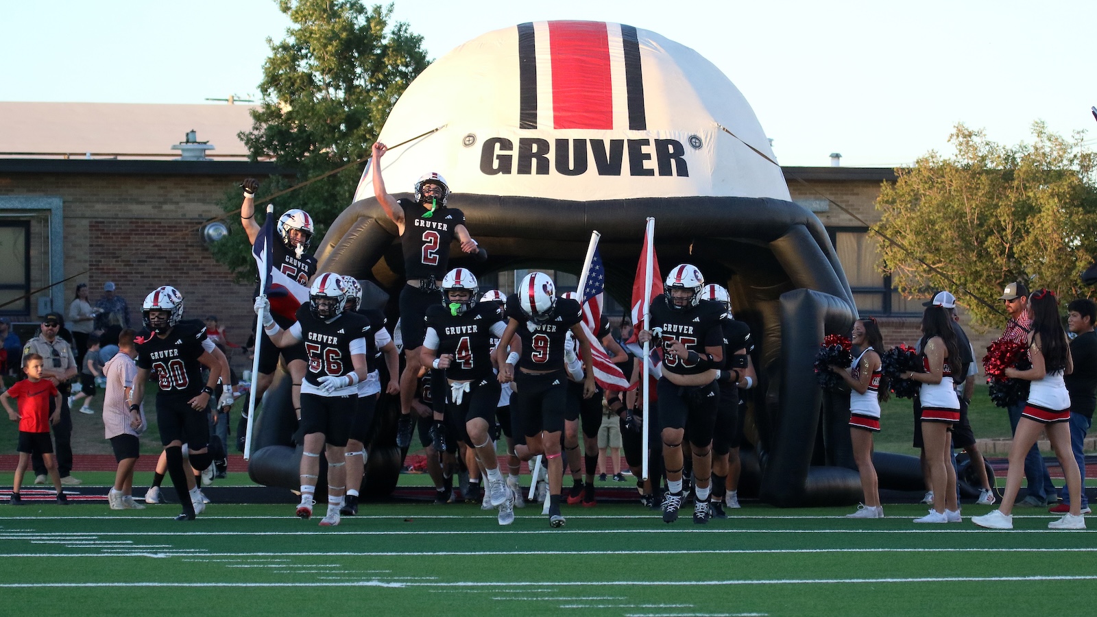 High school football team storms onto the field through a large inflatable Gruver helmet, cheerleaders on the sides.
