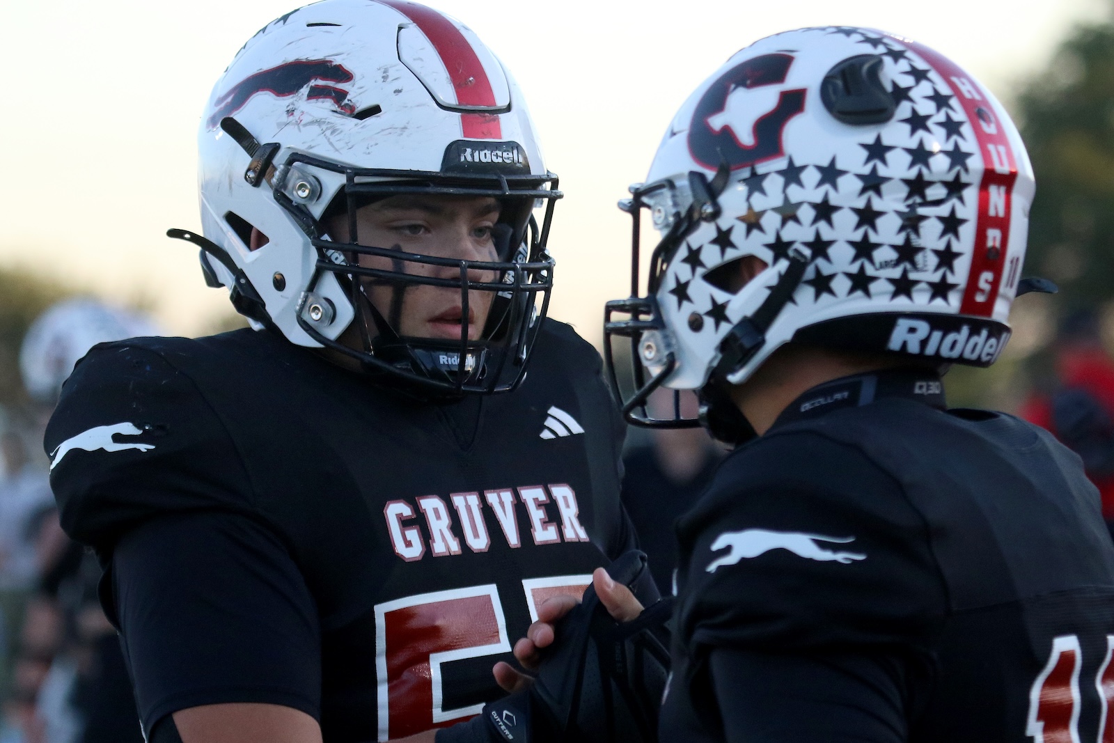 Two football players in black jerseys stand facing each other helmet-to-helmet on the field; one helmet is white with a red stripe while the other has a white star pattern.