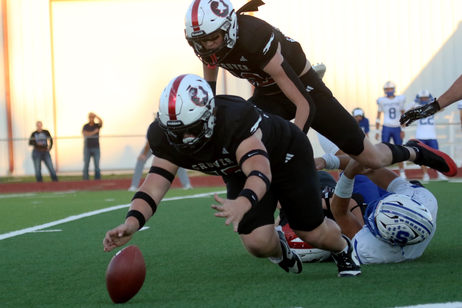 Football players in black uniforms dive for a loose ball on a green turf field, with white lines and opposing players in blue and white in the background.