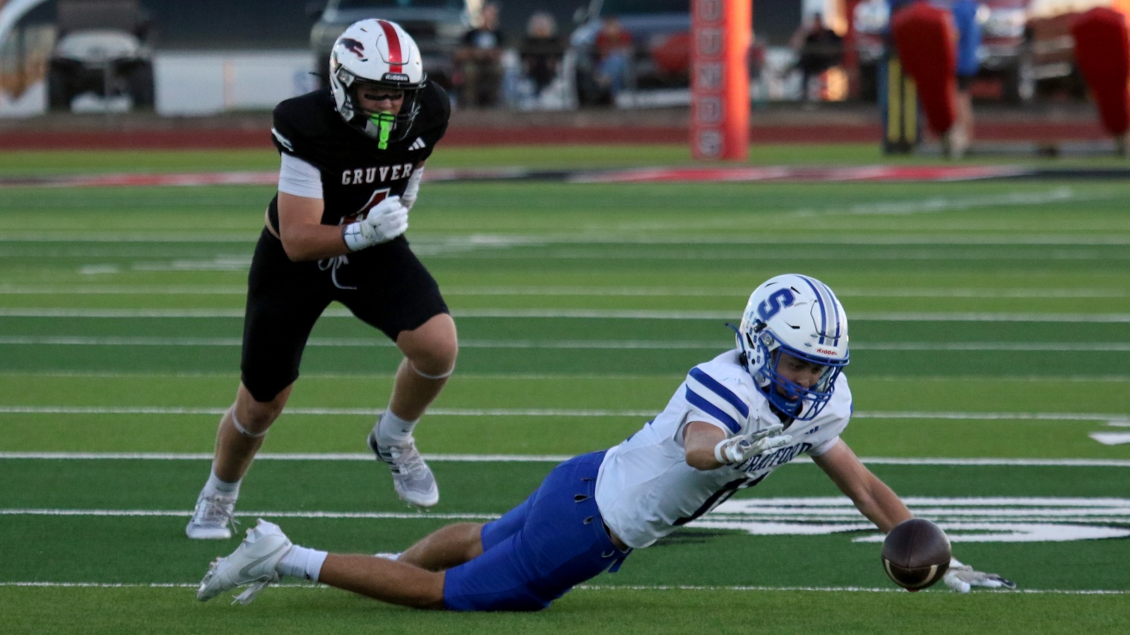 Football showdown: a white-and-blue jersey player dives on the turf reaching for the ball while a defender in black pursues nearby on a green field.