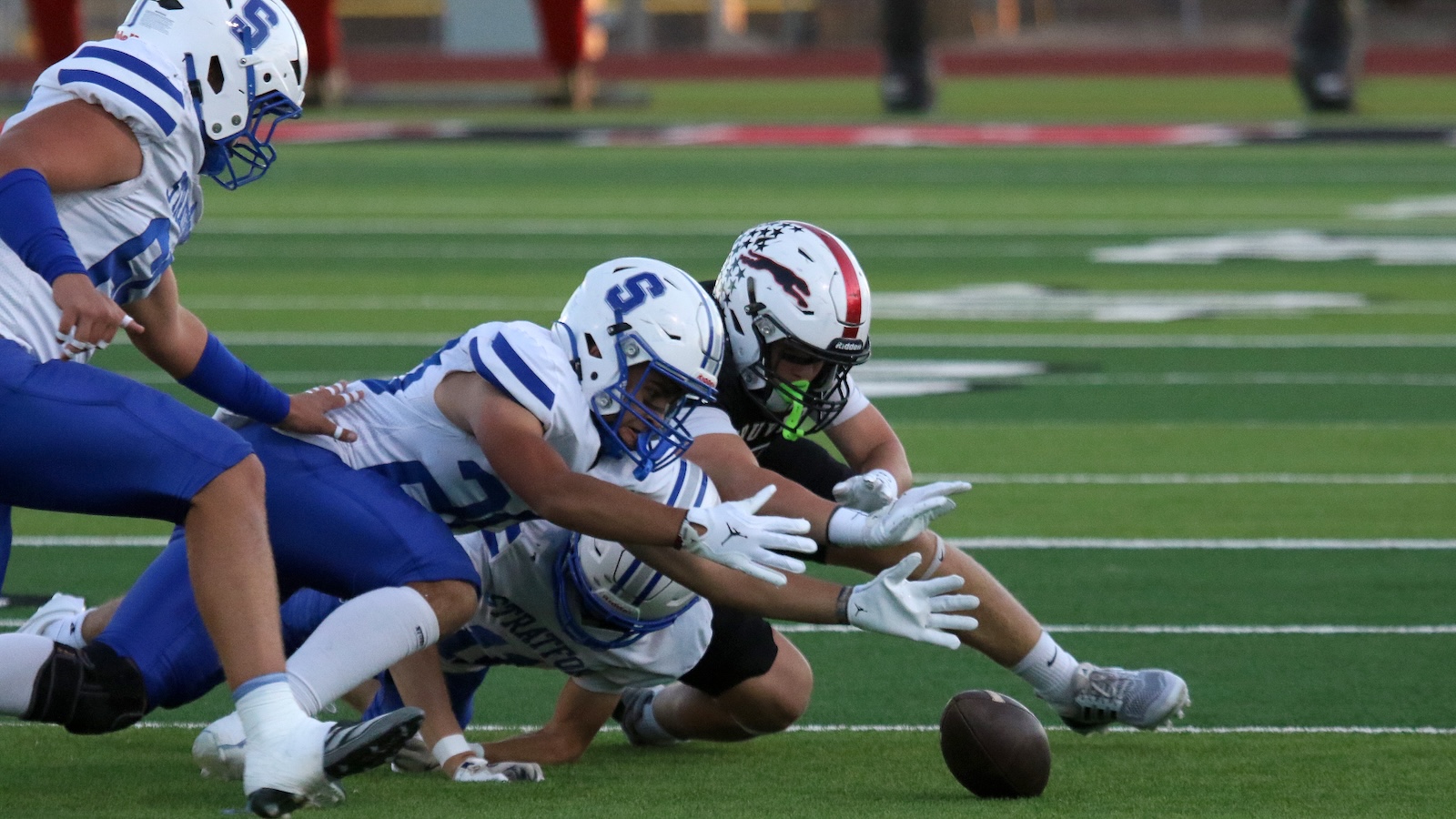 Football players in white and blue jerseys dive for a loose ball on a green field, helmets clashing as they reach for the ball near the ground, moment of a tackle in progress.