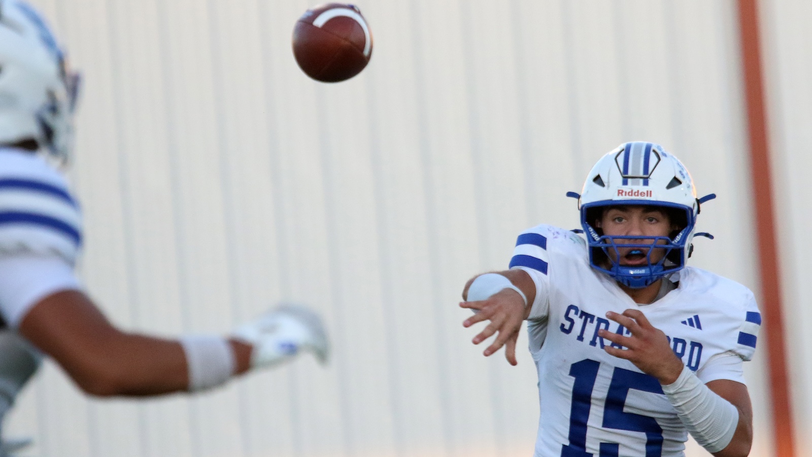 American football quarterback in a white and blue uniform throws a pass during a game, ball in midair near his outstretched hands .