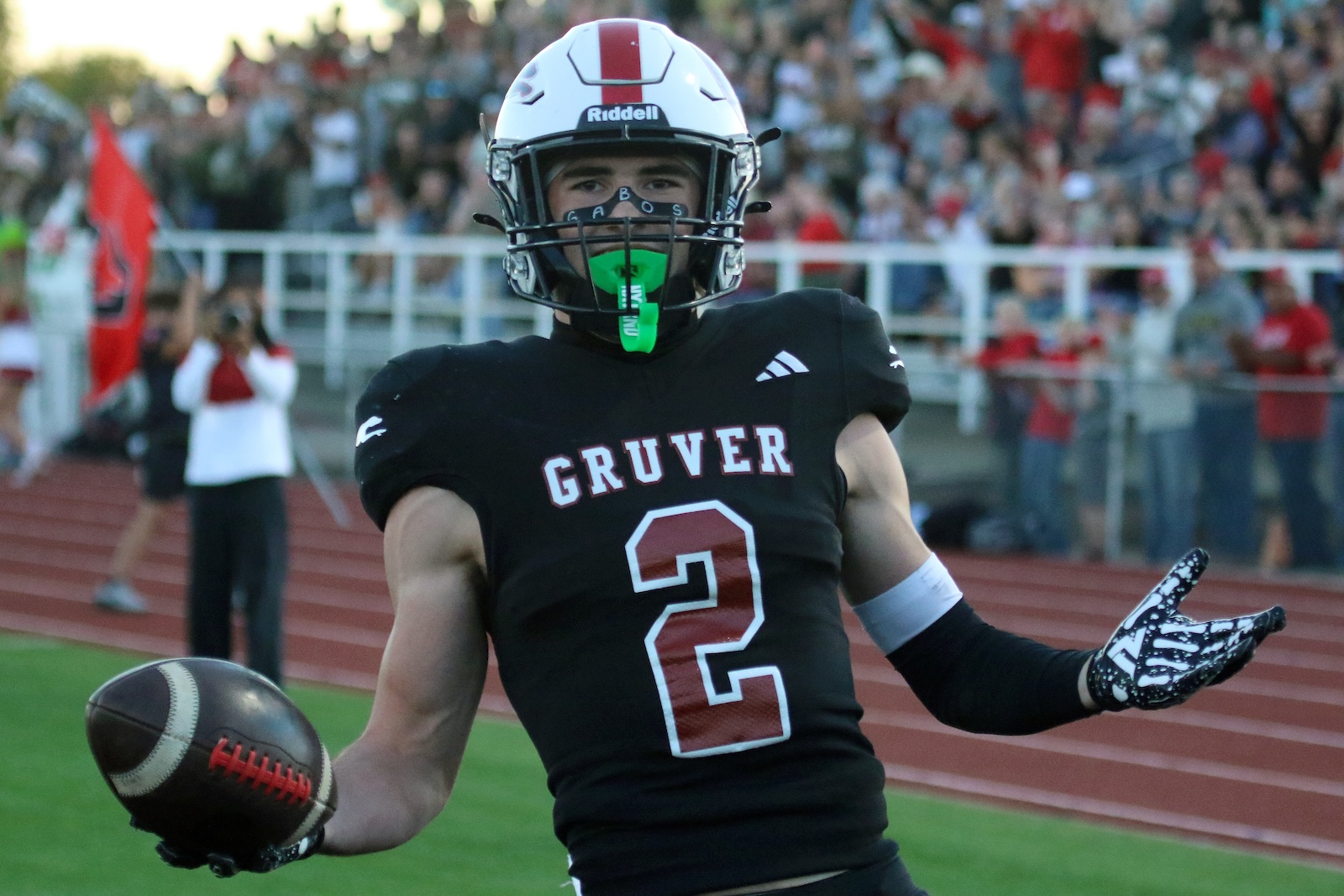Gruver football player wearing number 2 in a black jersey with a white helmet, holding a football and celebrating on a track with a crowd in the stands behind him.