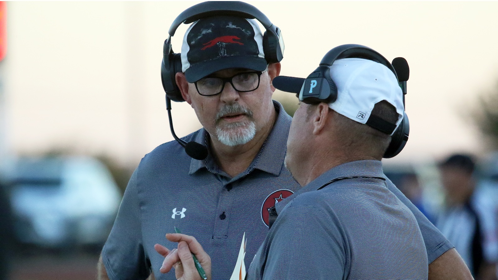 Two men in sports polos wearing headsets converse on the sideline, one holding papers or play notes.