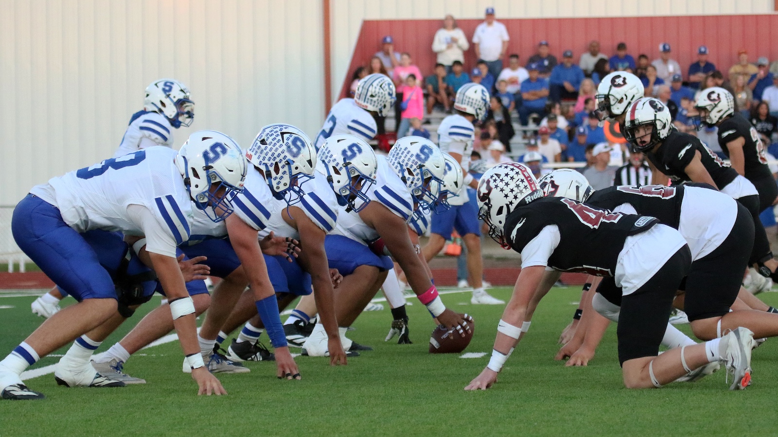 Football players line up at the line of scrimmage during a high school/college game, white-and-blue home team on offense vs. black-and-white visitors on defense on a green field with bleachers in the background