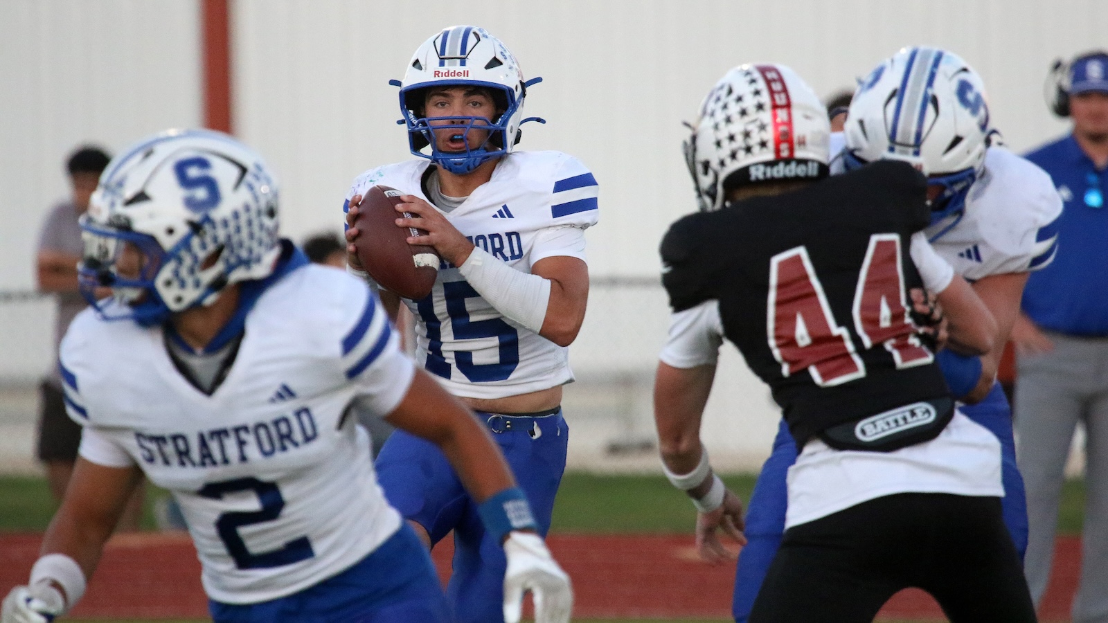 Stratford quarterback in white and blue holds the football, scanning for a pass while teammates block.