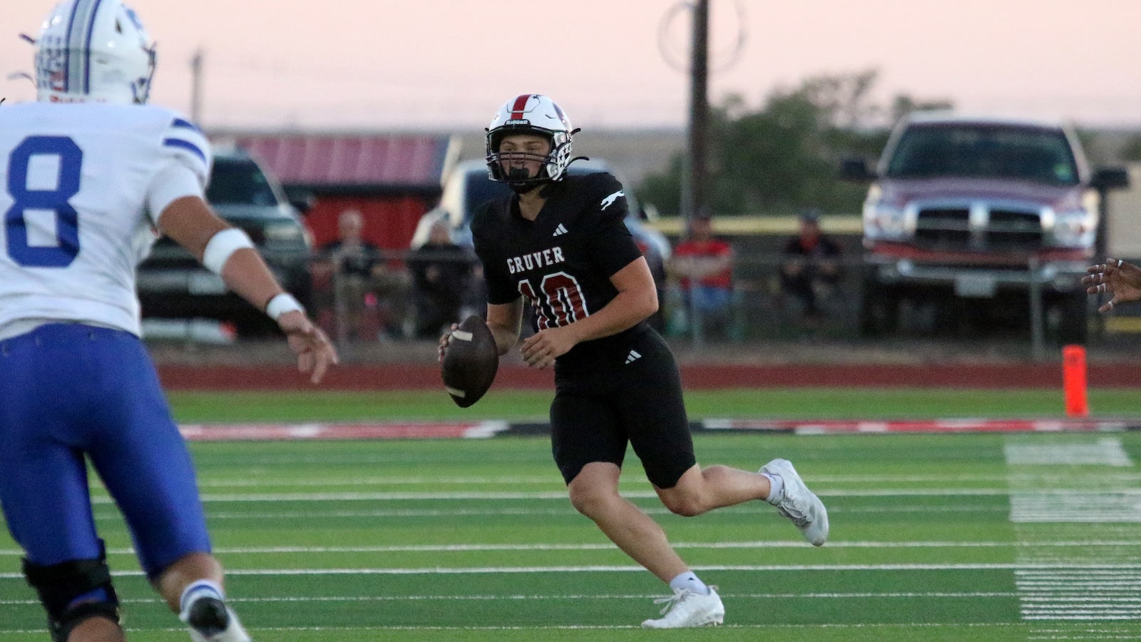 Gruver quarterback (number 10) in a black uniform runs with the football on a green turf field, pursued by a defender in white and blue.