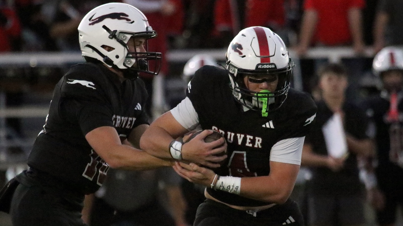 Two football players in black uniforms grapple for the ball on a game field, with a crowd in the background.