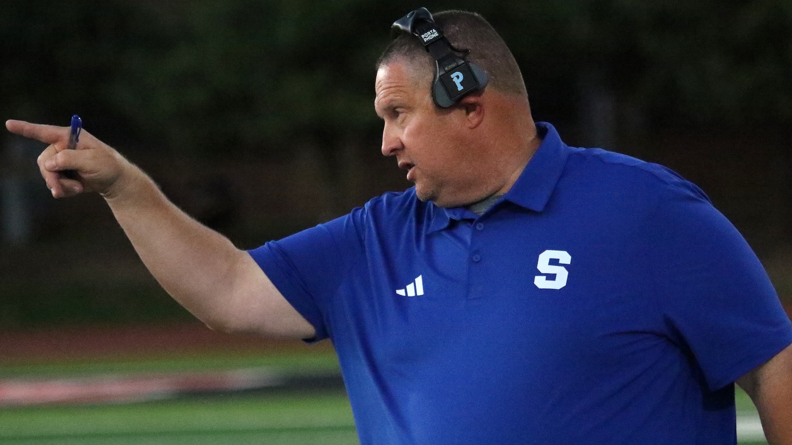 Coach in a blue polo points while wearing a headset on a sports field, directing players with a pen in hand
