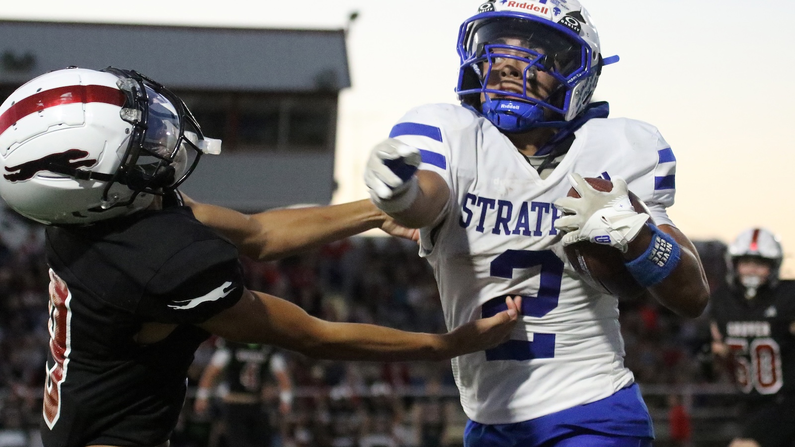 Football player in white and blue jersey reaches for a catch as a tackler in black closes in, during a high school/college game at dusk.